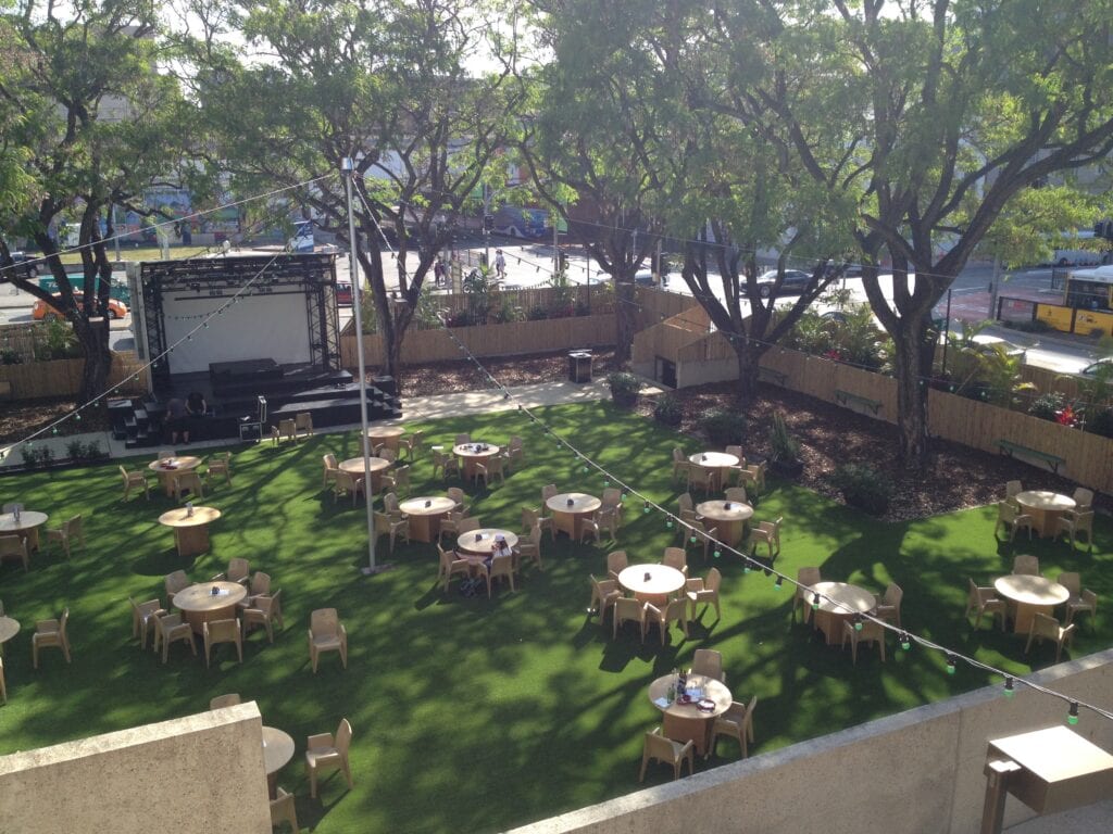 outdoor tables and chairs spread on fake grass with small music stage and backyard lighting