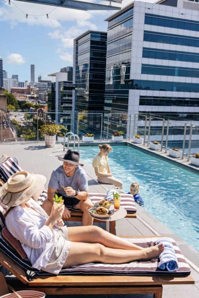 Couple enjoying lunch while kids swim in rooftop pool with city view